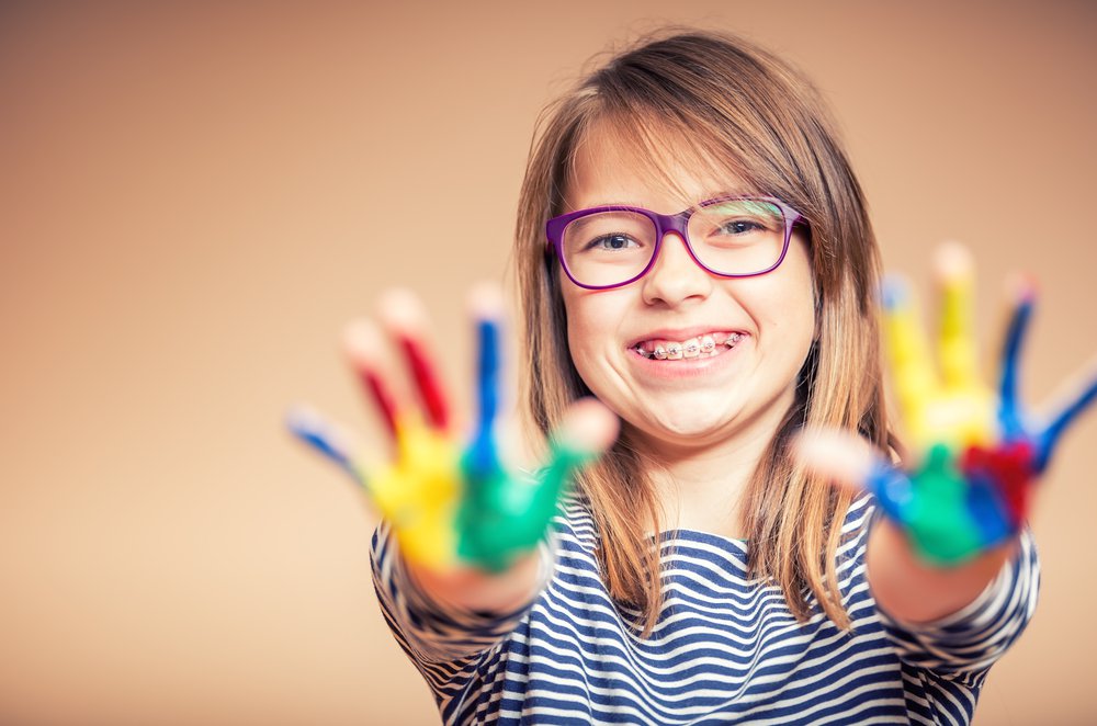 a happy young girl wearing braces holds up her hands to the camera showing they are covered in paint